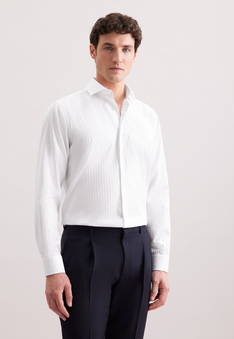 Camicia bianca a maniche lunghe con righe verticali, colletto button-down e polsini, abbinata a pantaloni eleganti blu navy. Texture del tessuto liscia.