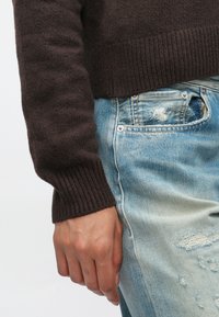 Χέρι ακουμπισμένο στο πλάι, φορώντας σκούρο καφέ πλεκτό πουλόβερ και ανοιχτό μπλε φθαρμένο τζιν με ορατή μπροστινή τσέπη και ραφές.