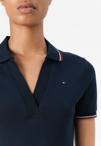 Tamsiai mėlyna polo marškinėliai su giliu V formos iškirpimu, su raudonai ir baltai dryžuotu apykakle ir mažu logotipo akcentu krūtinėje. Tekstūruota medžiaga.