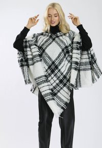 Poncho à carreaux noir et blanc en tissu doux, avec des bords frangés, porté sur un col roulé noir et un pantalon élégant.