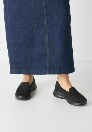 Black slip-on shoes with a textured mesh upper and cushioned sole, paired with a long denim skirt featuring a central seam.