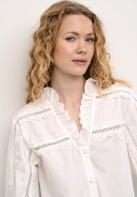 Balta blusas su ruffle apykakle ir nėrinių akcentais. Yra mygtukų uždarymas ir sudėtingi išpjovimo detalės ant rankovių ir krūtinės.