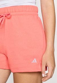 Calções atléticos corais com cintura elástica, cordão ajustável, bolsos laterais e um logótipo branco da Adidas na bainha inferior.