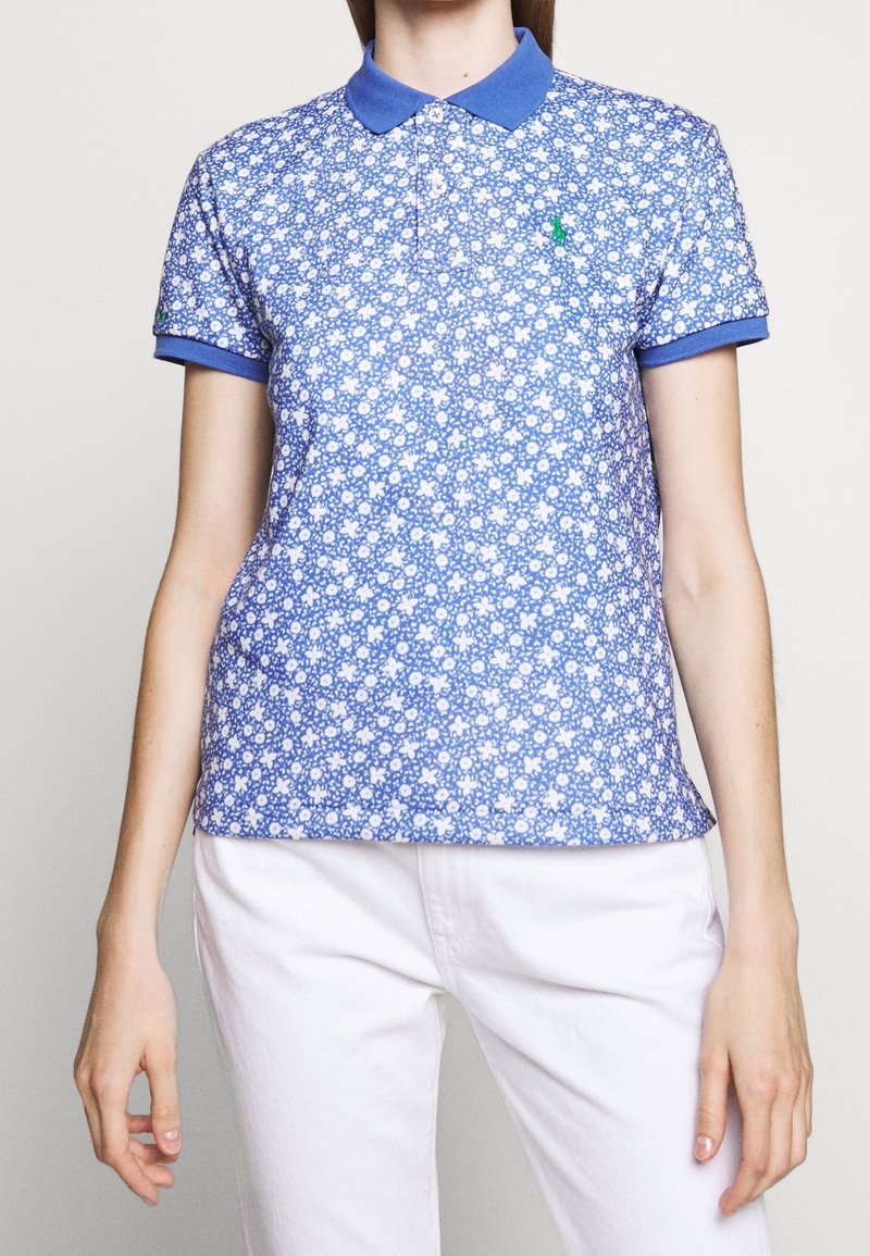 Femme portant un polo court bleu à motifs floraux avec un col bleu, associé à un pantalon blanc, debout devant un fond uni.