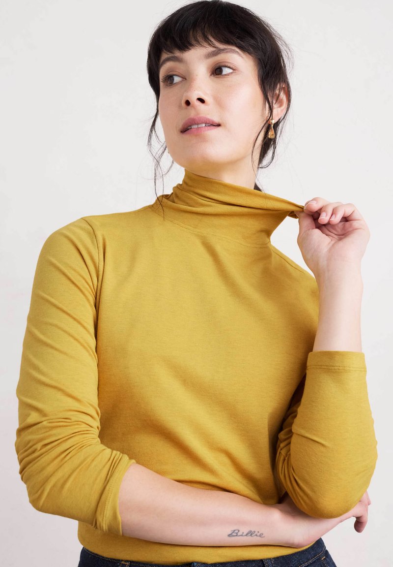 Sininen turtleneck-toppi pehmeästä kankaasta, jossa on istuvat pitkät hihat ja sileä pinta. Helma asettuu vyötärölle.