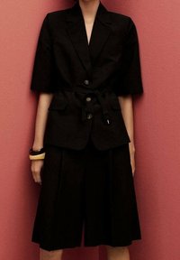 Czarna, dopasowana marynarka z krótkimi rękawami i paskiem, w połączeniu z czarnymi plisowanymi spodniami typu culottes. Gładka tekstura, z kołnierzykowym dekoltem i guzikami.
