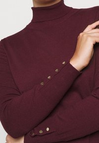 Pull à col roulé bordeaux avec texture côtelée, manches longues et boutons décoratifs dorés aux poignets. Conception douce et ajustée.