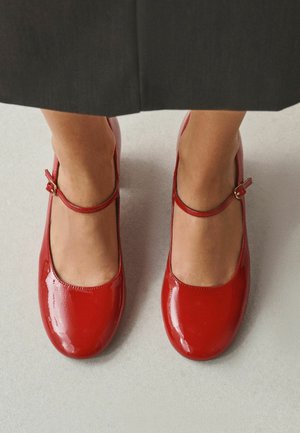 Feet wearing shiny red Mary Jane shoes with buckle straps, standing on a light-colored floor beneath a dark skirt hem.