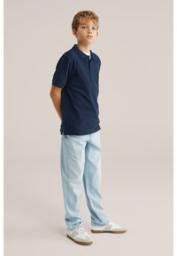 Poiss seisab käed selja taga, seljas tumelilla polo, heledad sinised teksad ja valged tossud, taust on ühtlane hele.