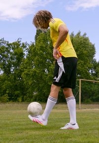Futballista sárga mezben, piros kiegészítőkkel, fekete nadrágban és fehér zokniban, ügyesen irányít egy fehér focilabdát a fűben.