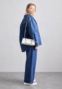 Nainen, jolla on yllään löysä sininen farkkutakki ja farkut, valkoinen käsilaukku ja valkoiset tennarit, seisoo sivuttain vaaleaa taustaa vasten.