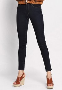 Donkerdenim skinny jeans met contrasterende oranje stiksels, voorzien van een middelhoge taille en vijf zakken. Gepaarde met bruine wedge sandalen.