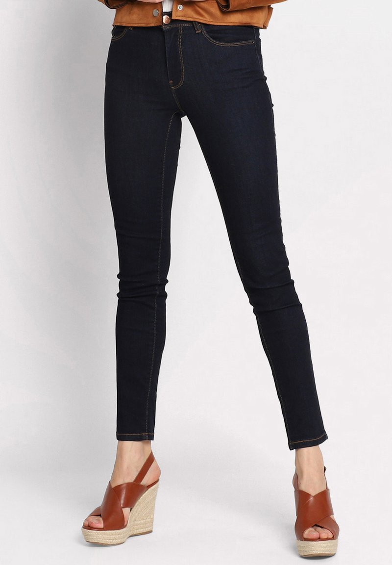 Donkerdenim skinny jeans met contrasterende oranje stiksels, voorzien van een middelhoge taille en vijf zakken. Gepaarde met bruine wedge sandalen.