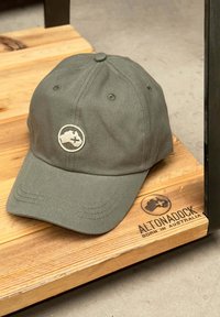 Gorra de béisbol color verde oliva hecha de tela texturizada, con un logotipo bordado en un parche circular en la parte frontal y visera curvada.