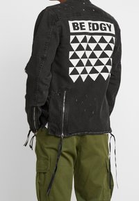 Henkilö, jolla on päällään musta farkkutakki, jossa on valkoisia geometrisia kolmioita ja selässä teksti "BE EDGY", sekä vihreät cargo-housut.