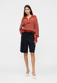 Γυναίκα με κόκκινο και άσπρο ριγέ πουλόβερ με κορδόνια στον γιακά, μαύρη βερμούδα μέχρι τα γόνατα και λευκές μυτερές γόβες, στέκεται μπροστά σε απλό φόντο.