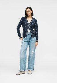 Μπλε ζακέτα ναυτικού με λευκή επένδυση και λουλουδάτο μοτίβο, συνδυασμένη με ανοιχτό μπλε τζιν παντελόνι με φαρδιά πόδια και μπεζ παπούτσια τύπου slip-on, φορεμένη πάνω από λευκό τοπ.