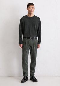 Mann in einem dunkelgrauen Langarmpullover, grünen Cordhosen mit schwarzem Gürtel und schwarzen Schnürschuhen steht vor einer weißen Wand.