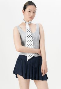 Mulher com cabelo escuro preso, vestindo uma blusa sem mangas de botões com riscas, uma saia azul com babados e um lenço de pescoço branco com bolinhas pretas, posando frente a um fundo branco.