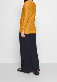 Senapsgul stickad tröja med diamantsmönster, långa ärmar och knapppynt, tillsammans med marinblå vida byxor.