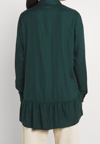 Kvinna i en mörkgrön långärmad blus med volangkant, sedd bakifrån, i kombination med ljusbeiga byxor.