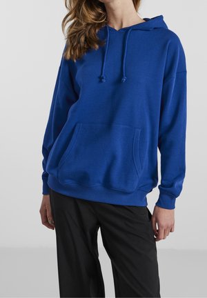 Mujer con sudadera azul con capucha, bolsillo delantero y cordones, combinada con pantalones negros holgados sobre un fondo claro liso.