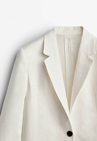 Valkoinen yksirivinen bleiseri, jossa on lovikannukset ja yksi näkyvä musta nappi vaalealla taustalla.