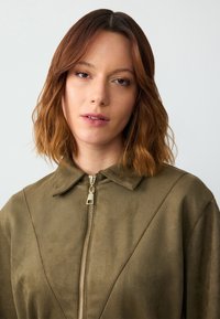 Giacca in suede verde oliva, morbida, con colletto a punta e chiusura frontale con zip in metallo dorato, cuciture dettagliate lungo le cuciture.