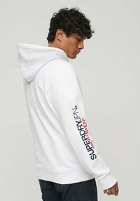 Felpa bianca con cappuccio, caratterizzata da un grande logo stampato in rosso e blu lungo la manica sinistra. Materiale morbido e vestibilità comoda.