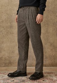 Trousers in a textured, brown fabric with a fine pattern, featuring front pleats and a tailored fit, paired with black leather shoes.