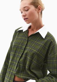 Camicia di flanella verde a quadri con colletto bianco, caratterizzata da una chiusura a bottoni frontale e maniche lunghe, che presenta una texture morbida e modelli a contrasto.