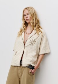 Cămașă bej cu mâneci scurte, cu nasturi, dintr-un material texturat, cu modele florale brodate și cusături contrastante. Asortată cu pantaloni kaki.