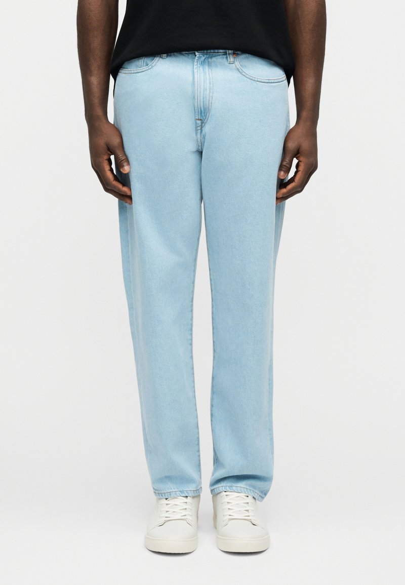 Άνδρας που φοράει ανοιχτό μπλε τζι ν με ίσια γραμμή, λευκά αθλητικά παπούτσια και μαύρο πουκάμισο, στέκεται μπροστά σε απλό λευκό φόντο.