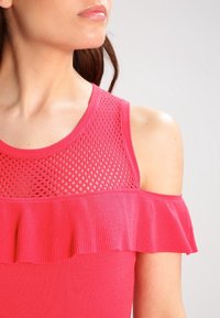 Top sans manches rose avec un design en maille au niveau du col et un accent en tissu volé le long du bord supérieur. Texture lisse et extensible.