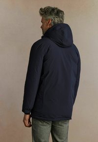 Granatowa kurtka z kapturem o gładkiej powłoce i elastycznych mankietach, z lekko wydłużonym tyłem dla dodatkowej osłony.