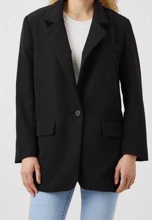 Femme portant un blazer noir à un seul bouton sur un top blanc côtelé et un jean bleu clair, debout devant un fond uni.