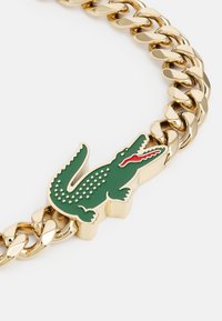 Goldene Kettenarmband mit einem grünen Krokodil-Logo und roten Akzenten, aus Metall, glatte Textur und verknüpftes Design.