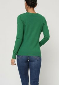 Pull en tricot vert à manches longues et ourlet côtelé, associé à un jean bleu. Le pull présente un design simple et une texture douce.