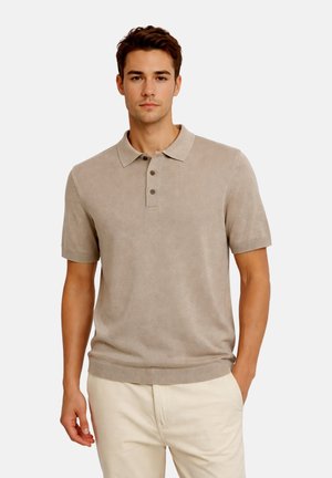 Giovane uomo che indossa una polo a maniche corte in maglia beige e pantaloni chiari, in piedi con una mano in tasca davanti a uno sfondo semplice.