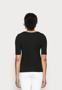 Person with curly hair wearing a black short-sleeve top and white pants, standing with back facing the viewer against a plain background.