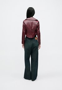 Hosszú fekete hajú nő bordó, rövid bőrkabátot és széles szárú sötét farmernadrágot visel, fehér, egyszerű háttér előtt áll.