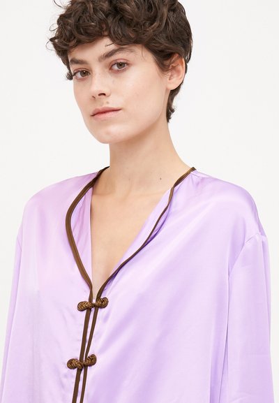 Top en soie lavande avec un décolleté en V profond, bordures marron et boutons décoratifs en nœud. Texture douce, coupe ample et design léger.
