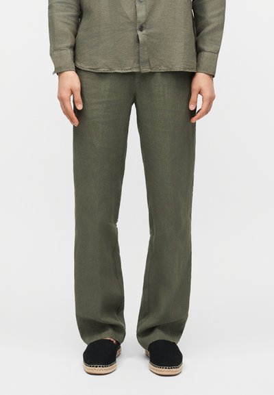 Homme portant un pantalon ample vert olive et une chemise boutonnée assortie avec des espadrilles noires sur un fond blanc uni.