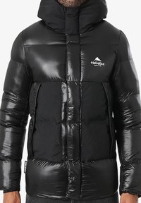 Doudoune noire à finition brillante, design matelassé, col montant et logo sur le côté gauche de la poitrine. Fermeture éclair à l'avant et poignets élastiques.