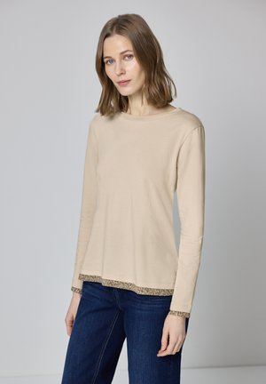 Beżowa bluzka z długim rękawem o zaokrąglonym dekolcie, z wzorzystym brzegiem i mankietami. Do tego dżinsy o luźnym kroju. Miękka faktura materiału.