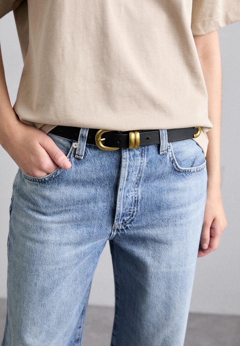 Svart skinnbälte med ett guldfärgat spänne, buren över ljusblå denimjeans. Skjortan är beige och tyget verkar mjukt och avslappnat.