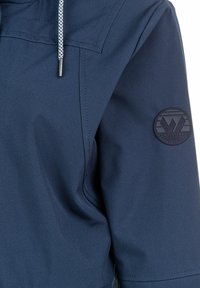 Navy blue jacket featuring a textured fabric, adjustable drawstring hood, and a circular black logo patch on the sleeve.