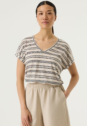 Haut à manches courtes en tissu léger avec un col en V, présentant une base beige ornée de motifs géométriques à rayures bleu foncé. Associé à un pantalon beige texturé.
