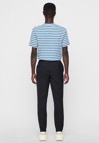 Muž stojící zády, oblečený v modro-bíle pruhovaném tričku s krátkým rukávem, černých kalhotách a bílých teniskách.