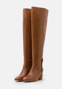 Bottes montantes en cuir marron à bout pointu, dotées d'un talon carré arrondi et d'un design minimaliste. Détail de liens noués à la cheville.